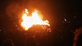 A large fire that is the focal point of an event taking place at night. A crowd of people is gathered in front of the flames, with shadows and silhouettes visible. The background is dark with a few small lights and green laser beams cutting across the scene.