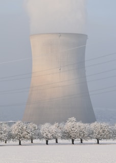 A cooling tower stands prominently in a snowy landscape. The tower emits steam into the overcast sky, with power lines crossing the scene. Snow-covered trees line the foreground, contrasting with the industrial structure.
