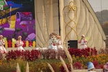 Four costume performers are on an outdoor stage, surrounded by vibrant flowers and greenery. The backdrop features a colorful, cartoon-style mural depicting a garden scene with a wheelbarrow, rake, and various plants. The performers are dressed in whimsical outfits, with one in the center wearing a character headpiece reminiscent of a mascot.