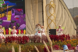 Four costume performers are on an outdoor stage, surrounded by vibrant flowers and greenery. The backdrop features a colorful, cartoon-style mural depicting a garden scene with a wheelbarrow, rake, and various plants. The performers are dressed in whimsical outfits, with one in the center wearing a character headpiece reminiscent of a mascot.