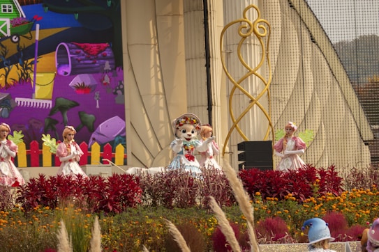 Four costume performers are on an outdoor stage, surrounded by vibrant flowers and greenery. The backdrop features a colorful, cartoon-style mural depicting a garden scene with a wheelbarrow, rake, and various plants. The performers are dressed in whimsical outfits, with one in the center wearing a character headpiece reminiscent of a mascot.