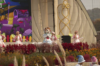 A colorful outdoor stage performance featuring mascots and performers in elaborate costumes. The central figure is a mascot wearing a blue dress and a floral headpiece, surrounded by dancers in white and pink outfits holding green leaf props. The backdrop includes a vibrant mural with abstract art and a whimsical design. The foreground is lush with red and orange flowers and ornamental grasses.