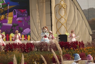 A colorful outdoor stage performance featuring mascots and performers in elaborate costumes. The central figure is a mascot wearing a blue dress and a floral headpiece, surrounded by dancers in white and pink outfits holding green leaf props. The backdrop includes a vibrant mural with abstract art and a whimsical design. The foreground is lush with red and orange flowers and ornamental grasses.