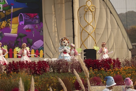 A colorful outdoor stage performance featuring mascots and performers in elaborate costumes. The central figure is a mascot wearing a blue dress and a floral headpiece, surrounded by dancers in white and pink outfits holding green leaf props. The backdrop includes a vibrant mural with abstract art and a whimsical design. The foreground is lush with red and orange flowers and ornamental grasses.