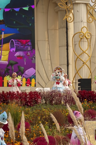 A colorful performance scene featuring characters in costumes standing on a stage. The backdrop includes vibrant, painted scenery. The area is adorned with a variety of flowering plants and ornamental grasses. The stage itself has decorative architectural elements.