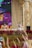 A colorful performance scene featuring characters in costumes standing on a stage. The backdrop includes vibrant, painted scenery. The area is adorned with a variety of flowering plants and ornamental grasses. The stage itself has decorative architectural elements.