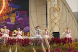 A group of performers dressed in colorful costumes stands on stage, surrounded by vibrant flowers and decorative elements. The setting includes a large backdrop with a whimsical illustration, contributing to a lively and festive atmosphere.