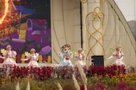 A group of performers dressed in colorful costumes stands on stage, surrounded by vibrant flowers and decorative elements. The setting includes a large backdrop with a whimsical illustration, contributing to a lively and festive atmosphere.