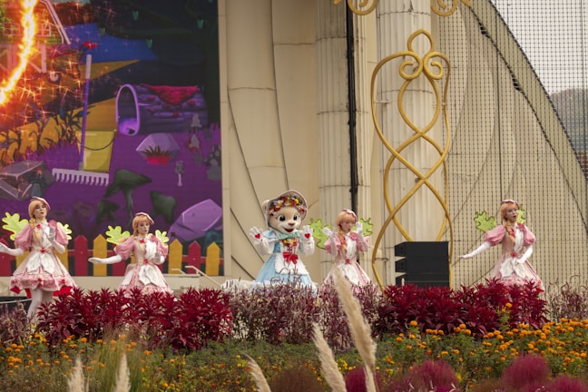 A group of performers dressed in colorful costumes stands on stage, surrounded by vibrant flowers and decorative elements. The setting includes a large backdrop with a whimsical illustration, contributing to a lively and festive atmosphere.
