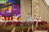 A group of costumed performers, including one in an animal suit, are on a decorated outdoor stage. Vibrant flowers and plants are in the foreground, with a backdrop depicting a colorful cartoon-like farm scene with a rake and various garden elements. The performers are in bright, whimsical outfits that complement the lively setting.