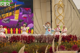 A group of costumed performers, including one in an animal suit, are on a decorated outdoor stage. Vibrant flowers and plants are in the foreground, with a backdrop depicting a colorful cartoon-like farm scene with a rake and various garden elements. The performers are in bright, whimsical outfits that complement the lively setting.