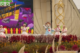 A group of costumed performers, including one in an animal suit, are on a decorated outdoor stage. Vibrant flowers and plants are in the foreground, with a backdrop depicting a colorful cartoon-like farm scene with a rake and various garden elements. The performers are in bright, whimsical outfits that complement the lively setting.