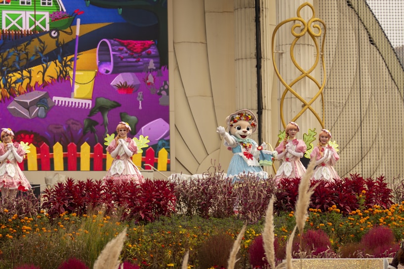 A group of costumed performers, including one in an animal suit, are on a decorated outdoor stage. Vibrant flowers and plants are in the foreground, with a backdrop depicting a colorful cartoon-like farm scene with a rake and various garden elements. The performers are in bright, whimsical outfits that complement the lively setting.