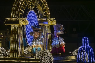 Two costumed characters lit up by numerous colorful lights stand on a decorative float. The scene is set during nighttime, with the characters surrounded by sparkling lights and reflective surfaces. A backdrop of structures lighting makes the atmosphere festive.