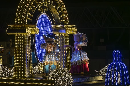 Two costumed characters lit up by numerous colorful lights stand on a decorative float. The scene is set during nighttime, with the characters surrounded by sparkling lights and reflective surfaces. A backdrop of structures lighting makes the atmosphere festive.