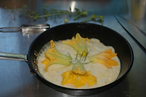 A frying pan containing a creamy mixture with yellow zucchini flowers laid on top, positioned on a metal surface. In the background, there is a peeler, herbs, and a knife.