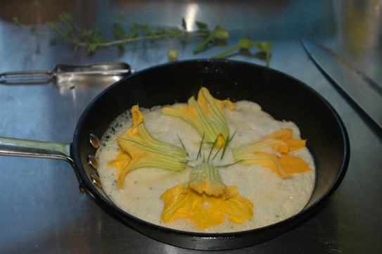 A frying pan containing a creamy mixture with yellow zucchini flowers laid on top, positioned on a metal surface. In the background, there is a peeler, herbs, and a knife.
