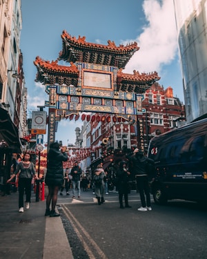 A bustling Chinatown street scene with a prominent traditional Chinese archway adorned in red, blue, and gold colors. The street is filled with people, including a woman taking a photo and others walking or talking. Red lanterns are strung across the street, and the setting is surrounded by buildings with red brick facades.