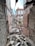 Rubble and debris are scattered across the floor of a dilapidated, roofless structure with crumbling brick walls. Vegetation is visible through broken window frames, hinting at neglect and decay. The building appears abandoned, with damaged beams and exposed surfaces.