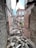 Rubble and debris are scattered across the floor of a dilapidated, roofless structure with crumbling brick walls. Vegetation is visible through broken window frames, hinting at neglect and decay. The building appears abandoned, with damaged beams and exposed surfaces.