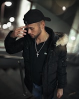 A man is wearing a black cap backwards and a black coat with a fur-trimmed hood. He is looking downwards with his hand raised to his forehead, wearing a cross necklace. The background is blurred with circular lights, possibly indicating an urban setting at night.