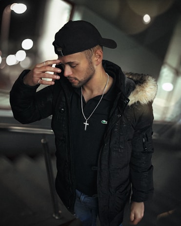 A man is wearing a black cap backwards and a black coat with a fur-trimmed hood. He is looking downwards with his hand raised to his forehead, wearing a cross necklace. The background is blurred with circular lights, possibly indicating an urban setting at night.