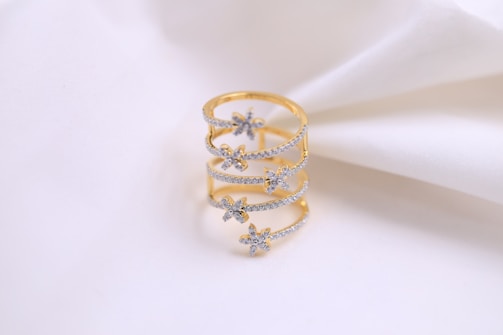 A gold ring with delicate floral designs encrusted with small diamonds, displayed on a soft white fabric background.