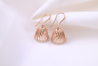 A pair of elegant rose gold earrings with a delicate, flowing design. The earrings have a cage-like structure embellished with small, sparkling gemstones at the bottom. They are displayed on a soft, white fabric background, enhancing their luxurious appearance.