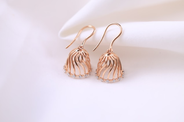 A pair of elegant rose gold earrings with a delicate, flowing design. The earrings have a cage-like structure embellished with small, sparkling gemstones at the bottom. They are displayed on a soft, white fabric background, enhancing their luxurious appearance.