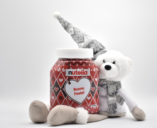 A large jar of Nutella with festive red and white packaging. The jar features a heart design with the message 'Buone Feste!' written on it. It's accompanied by a white plush bear wearing a patterned gray hat and scarf, suggesting a holiday theme.