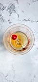 A vintage-style glass with ornate patterns contains a cocktail with a maraschino cherry on a stick. It rests on a white marble surface with grey veining.