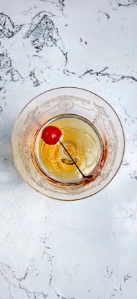 A vintage-style glass with ornate patterns contains a cocktail with a maraschino cherry on a stick. It rests on a white marble surface with grey veining.