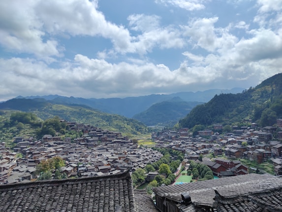 A village with traditional-style wooden houses nestled in a lush, mountainous landscape. The view features densely clustered rooftops and is framed by verdant hills under a partly cloudy sky.