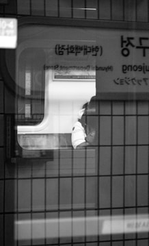 A black and white photograph capturing the interior of a train, viewed through a window marked with Korean text. Inside the train, a person in a light-colored jacket is sitting alone, partially obscured by the window frame. The seats and wall of the train are also visible, lending a sense of quiet isolation.