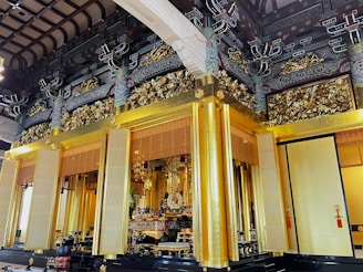 An ornate temple interior featuring intricate woodwork and lavish gold decorations. The walls and ceiling are adorned with detailed carvings and patterns, while the space displays religious offerings and sacred symbols.