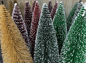A collection of decorative Christmas trees with textured surfaces and snow-like accents. The trees vary in colors including gold, red, and green, and are densely packed together.