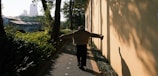 A person walks along a narrow pathway bordered by lush greenery on one side and a sunlit wall on the other. The individual has their arms outstretched and is casting a shadow on the wall. Tall trees and urban buildings are visible in the background, partially obscured by foliage.