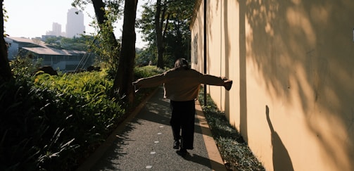 A person walks along a narrow pathway bordered by lush greenery on one side and a sunlit wall on the other. The individual has their arms outstretched and is casting a shadow on the wall. Tall trees and urban buildings are visible in the background, partially obscured by foliage.