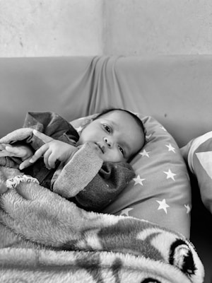 An infant lying on a cushioned surface, partially covered with a soft, patterned blanket. The baby is wearing a dark onesie and is surrounded by a pillow with a star pattern. The setting appears calm and comfortable.