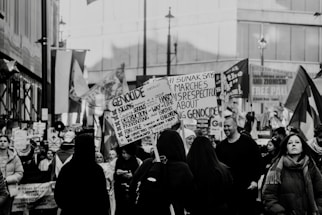 A black and white image of a street protest or demonstration, where a crowd of people is gathered holding various signs and banners related to political or social issues. The setting appears to be an urban area with buildings in the background. The crowd consists of diverse individuals, some of whom are engaged in reading or holding up placards with printed and handwritten messages.