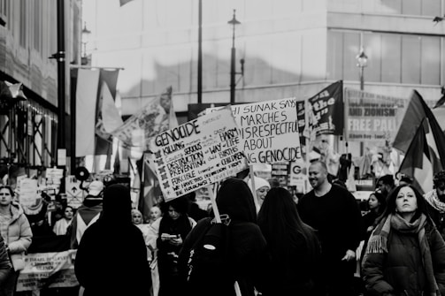 A black and white image of a street protest or demonstration, where a crowd of people is gathered holding various signs and banners related to political or social issues. The setting appears to be an urban area with buildings in the background. The crowd consists of diverse individuals, some of whom are engaged in reading or holding up placards with printed and handwritten messages.