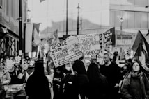 A black and white image of a street protest or demonstration, where a crowd of people is gathered holding various signs and banners related to political or social issues. The setting appears to be an urban area with buildings in the background. The crowd consists of diverse individuals, some of whom are engaged in reading or holding up placards with printed and handwritten messages.