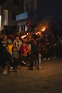 A street performer entertains a crowd with fiery torches at night. The audience, consisting of children and adults, are gathered closely around the performer, watching attentively. The scene is illuminated by the glow of the flames, casting warm light on the spectators. The background shows a dimly lit urban setting with buildings and signs.