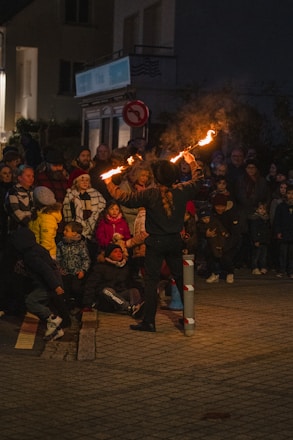 A street performer entertains a crowd with fiery torches at night. The audience, consisting of children and adults, are gathered closely around the performer, watching attentively. The scene is illuminated by the glow of the flames, casting warm light on the spectators. The background shows a dimly lit urban setting with buildings and signs.