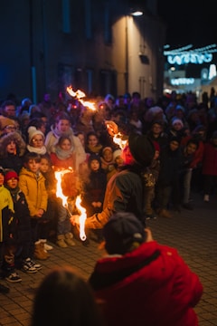 A performer is juggling with flaming torches in front of a crowd of spectators, including many children. The scene takes place outdoors, likely during a festival or public event at night. The audience is bundled up in winter clothing, suggesting cold weather.