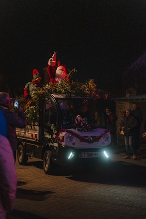 A festive scene features a small vehicle adorned with holiday decorations, including greenery and red tinsel. On top of the vehicle, a person dressed as Santa Claus, complete with a red hat and white beard, is waving or gesturing. Next to Santa stands another person wearing an elf costume. The scene takes place at night, surrounded by dark skies with a few onlookers visible around the vehicle, capturing the moment with their phones.