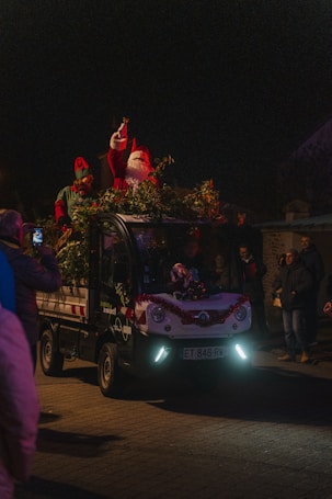 A festive scene features a small vehicle adorned with holiday decorations, including greenery and red tinsel. On top of the vehicle, a person dressed as Santa Claus, complete with a red hat and white beard, is waving or gesturing. Next to Santa stands another person wearing an elf costume. The scene takes place at night, surrounded by dark skies with a few onlookers visible around the vehicle, capturing the moment with their phones.