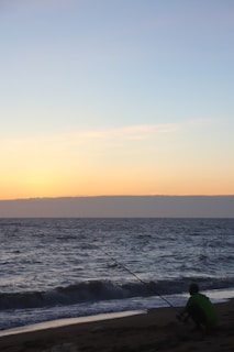 A person is fishing on a sandy beach at sunrise or sunset, with a calm ocean and clear sky. The horizon is visible, and a gentle wave is approaching the shore.