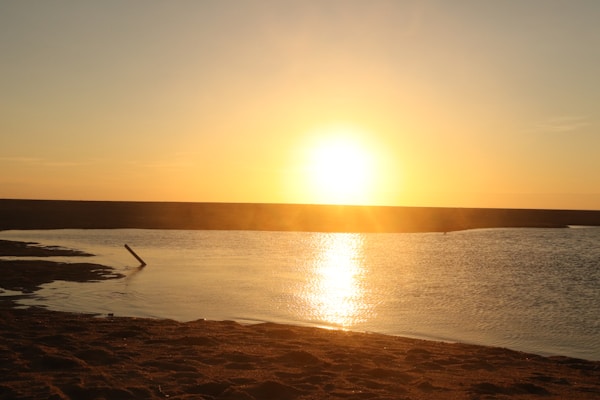 The horizon shows the sun setting over a body of water, creating a warm glow that reflects off the surface. The foreground includes a sandy shore with gentle waves, and the color palette is dominated by warm hues.
