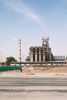 An industrial facility with large cylindrical metal structures and a tall chimney that has red and white stripes. The foreground shows a person in an orange vest working with a tripod and surveying equipment. The sky is clear with a few clouds, and the area around the facility appears under construction or renovation.